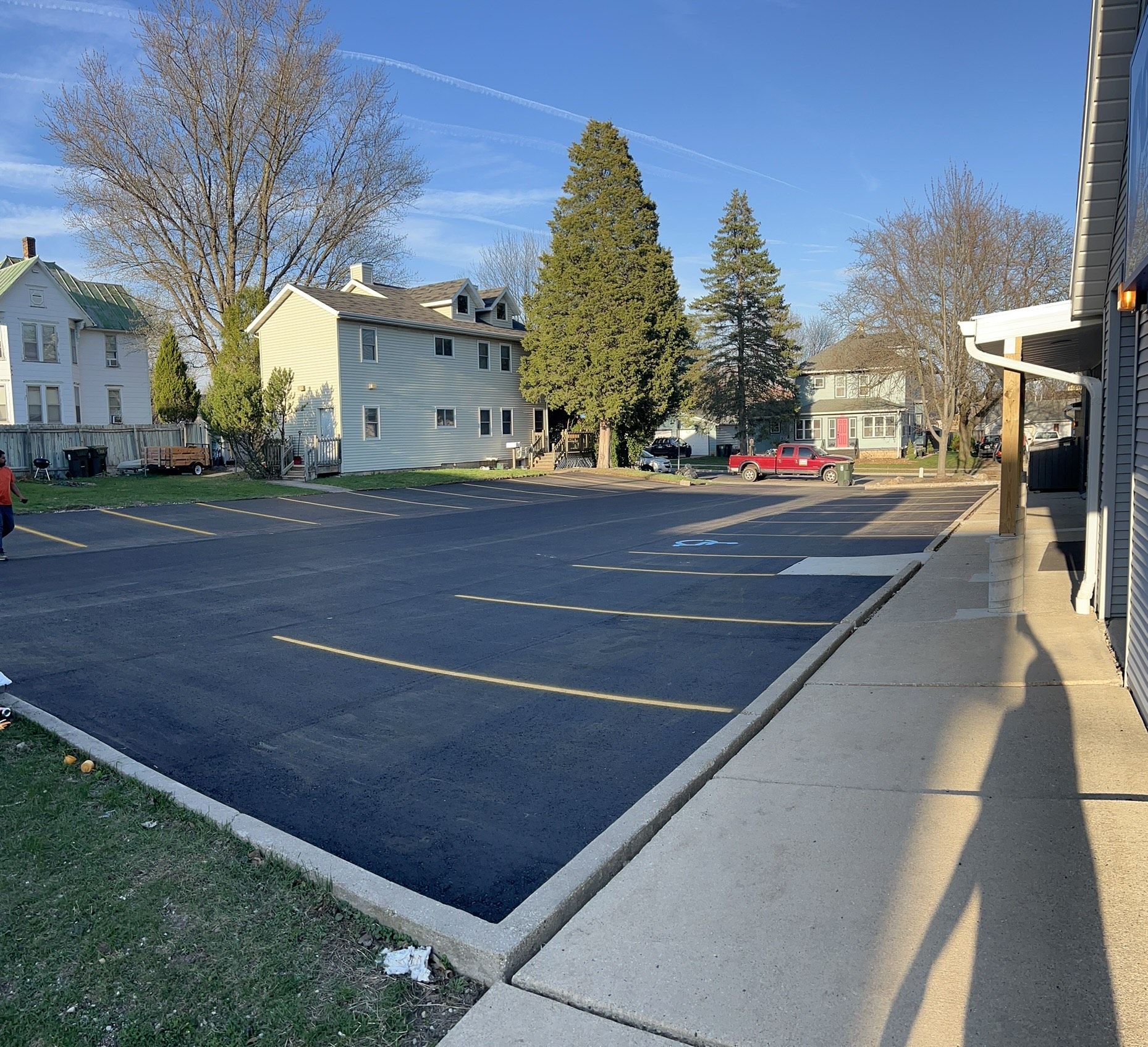 Paved parking lot with yellow lines, bordered by a sidewalk and buildings under a blue sky.