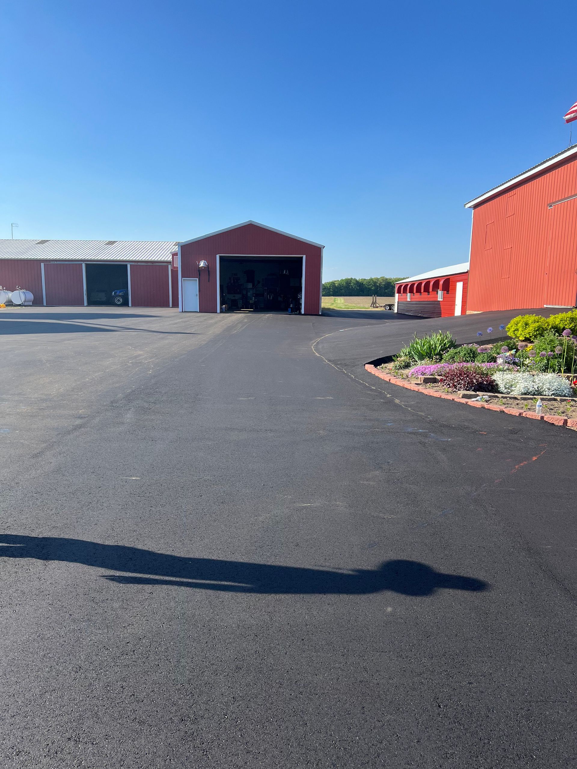 Paved driveway leads to red barns under a bright blue sky; flower bed on the right.
