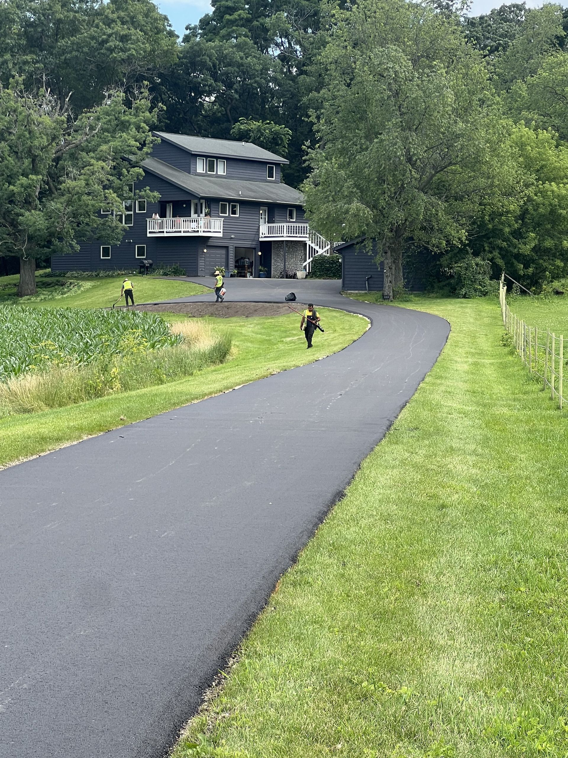 Black asphalt driveway winds toward a gray house; workers tend to the lawn.