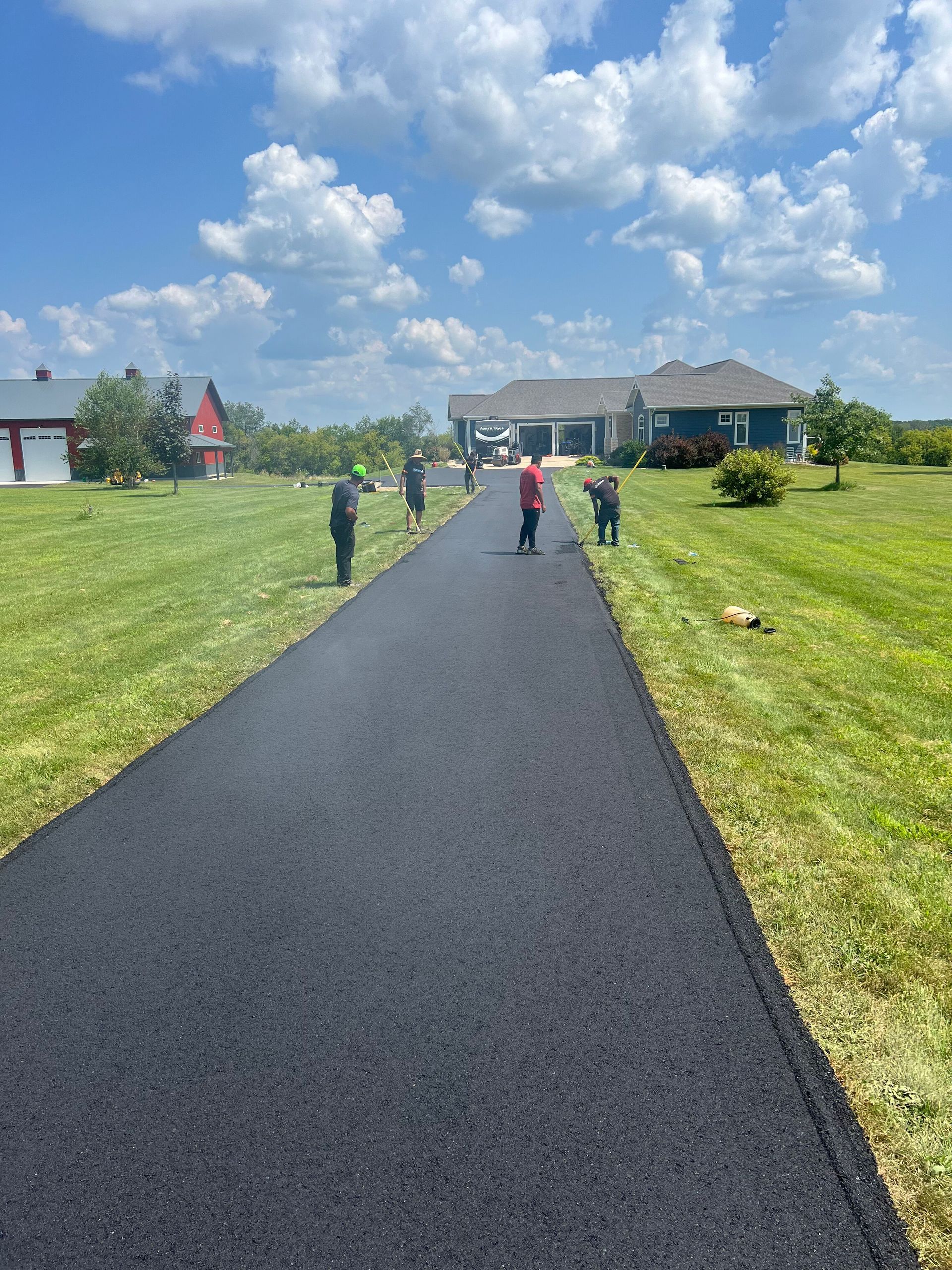 Workers paving a long asphalt driveway towards a house on a sunny day.