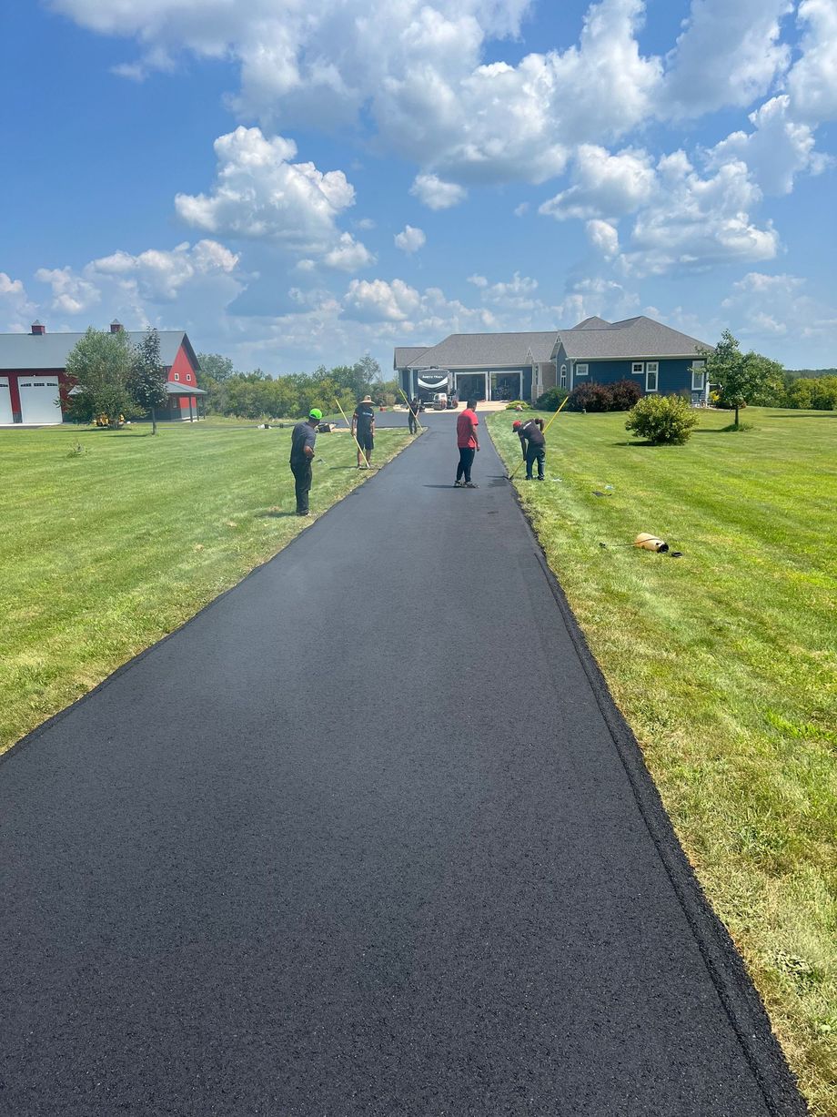 Workers paving a long asphalt driveway towards a house on a sunny day.