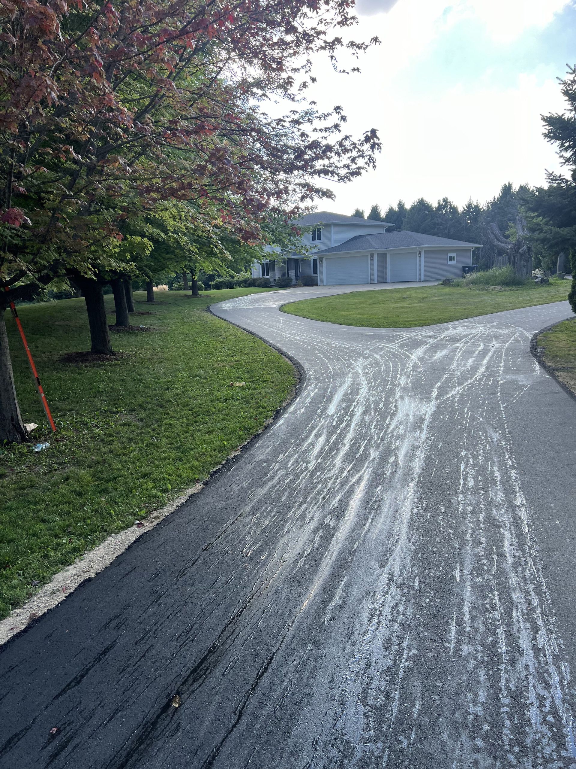 Asphalt driveway splits, leading to a house with a garage, trees on the left, and grass in the middle.