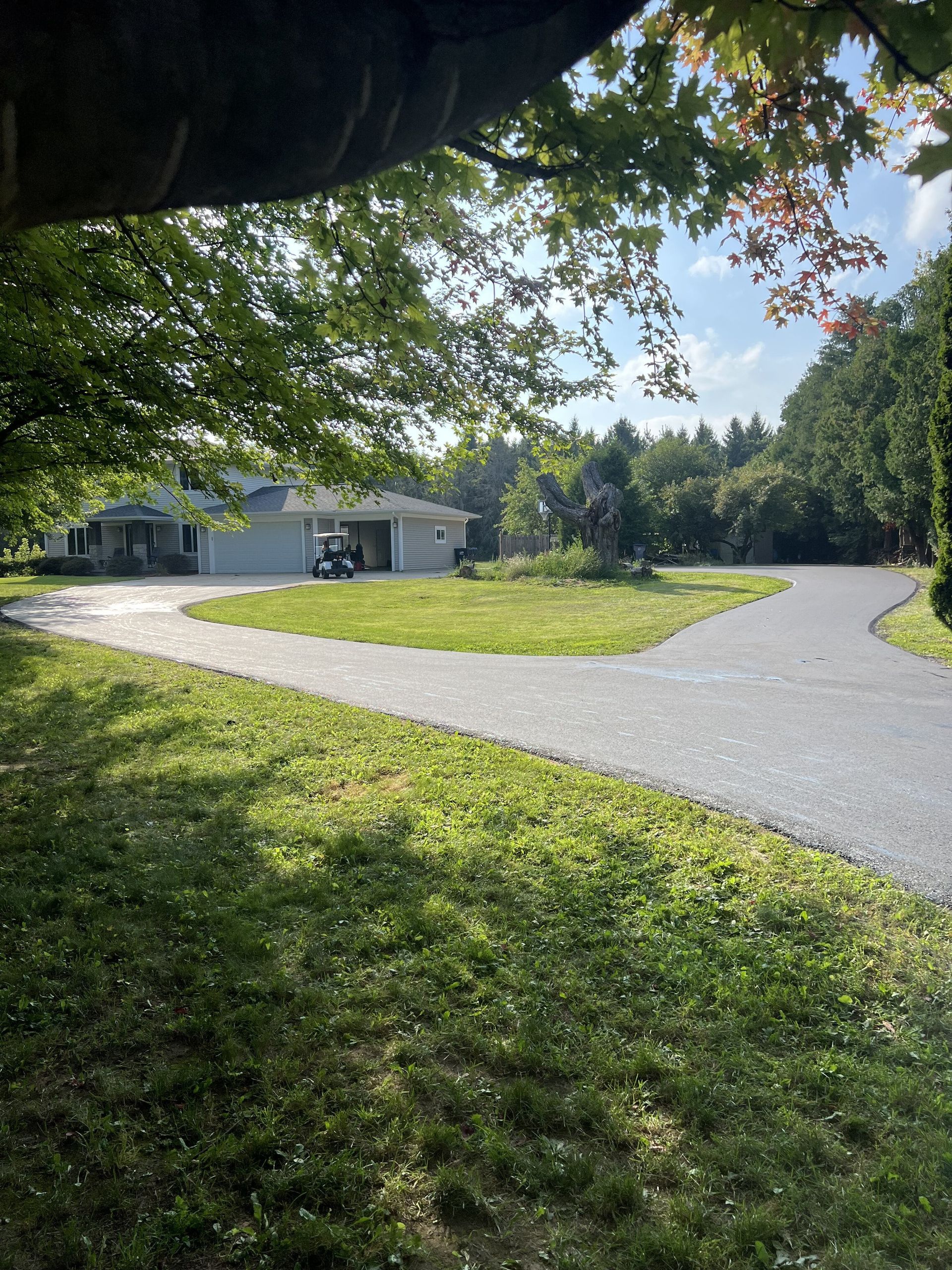 Paved driveway leading to a light-colored house with a golf cart, surrounded by green grass and trees.