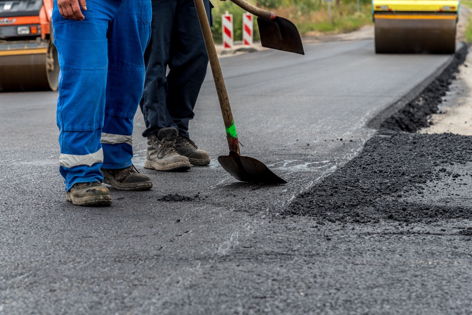 Road workers paving asphalt roadway with shovels and roller during road construction repairs project.