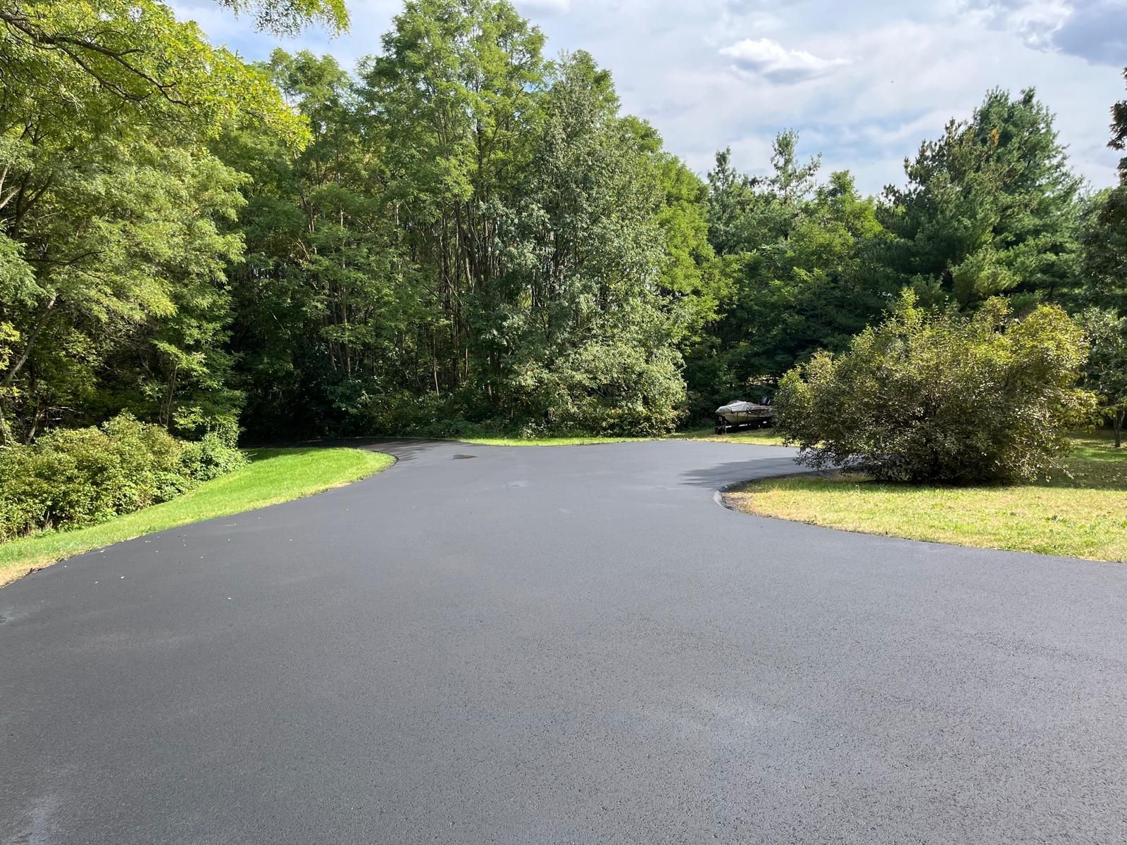 Newly paved black asphalt driveway curves into a wooded area with green grass and trees.