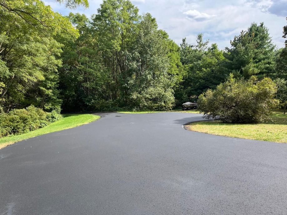 Newly paved black asphalt driveway curves into a wooded area with green grass and trees.