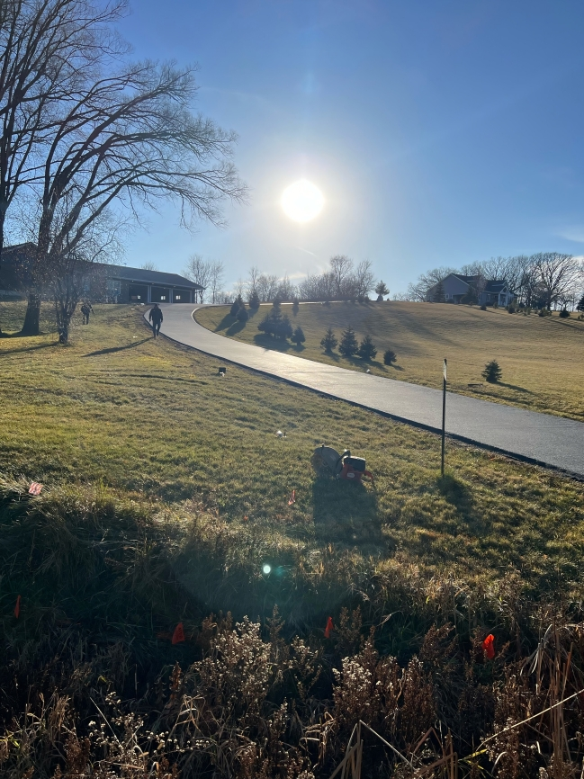 A sunny outdoor scene: a paved road curves uphill through a grassy field towards a house under a bright sun.