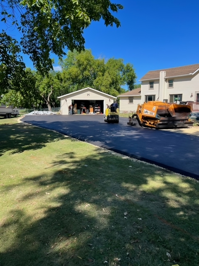 Paved driveway in front of a garage and a house, with construction equipment and workers, on a sunny day.