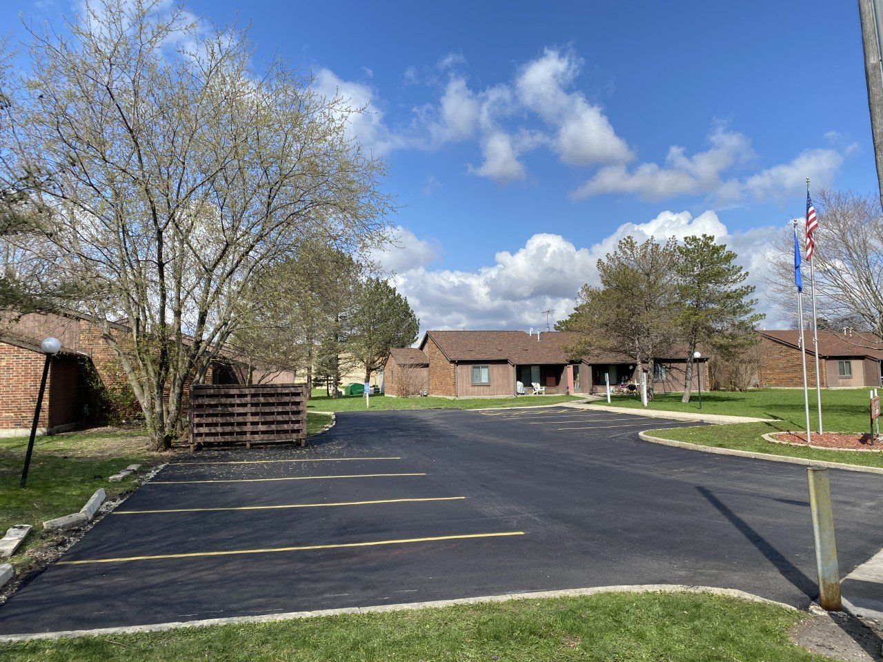 Paved parking area in front of a low-rise building under a blue sky with clouds. Two flags fly in the distance.