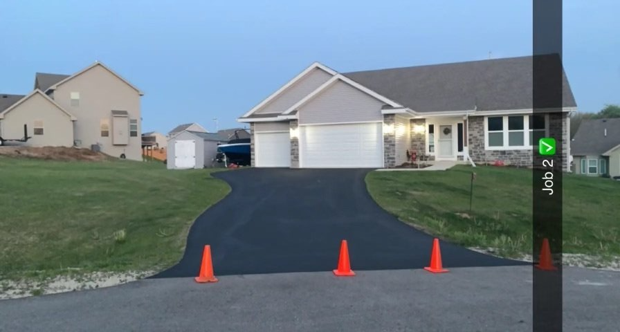 A newly paved asphalt driveway leading to a suburban house; orange cones are in the drive.
