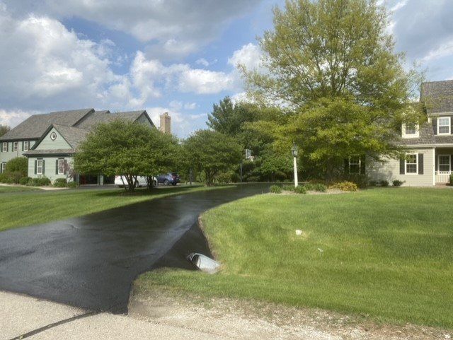 Paved road curves through a residential neighborhood with green lawns and trees on a sunny day.