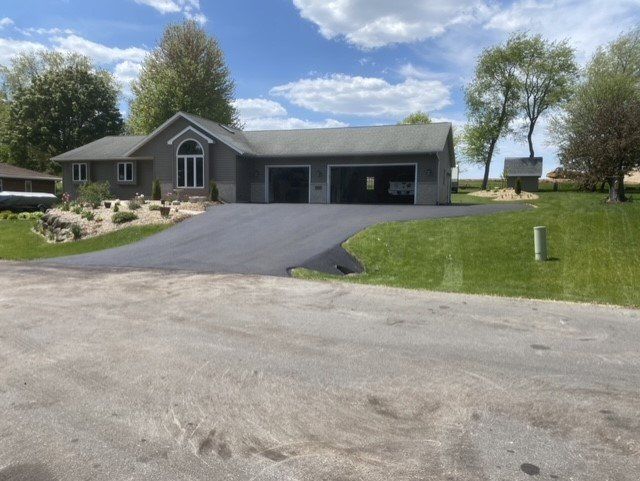 A gray house with a two-car garage and a paved driveway, surrounded by green grass and blue sky.