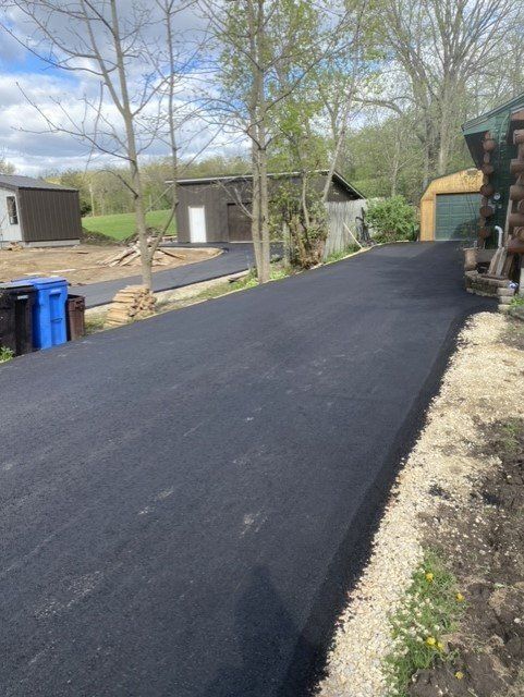 Newly paved asphalt driveway lined with gravel, leading toward small buildings and trees.
