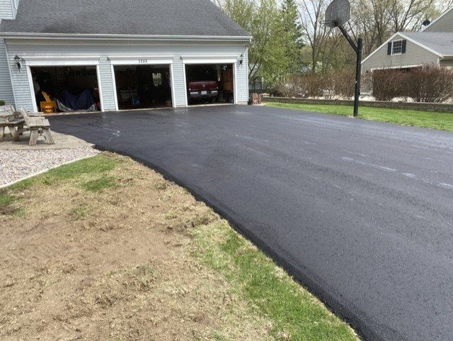 Black asphalt driveway leading to a three-car garage, with green grass border, a basketball hoop, and a house in the background.