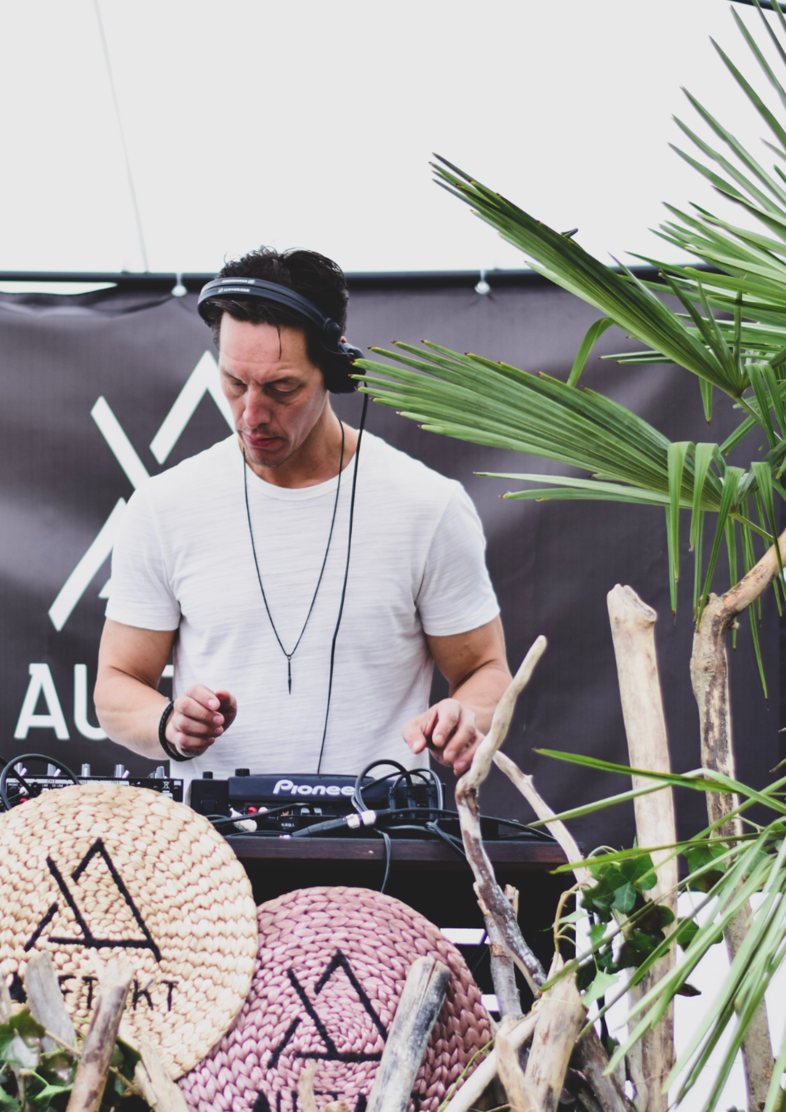 Ein DJ mit Kopfhörern und weißem T-Shirt legt bei einer mit Pflanzen geschmückten Open-Air-Veranstaltung hinter einem Mischpult auf.