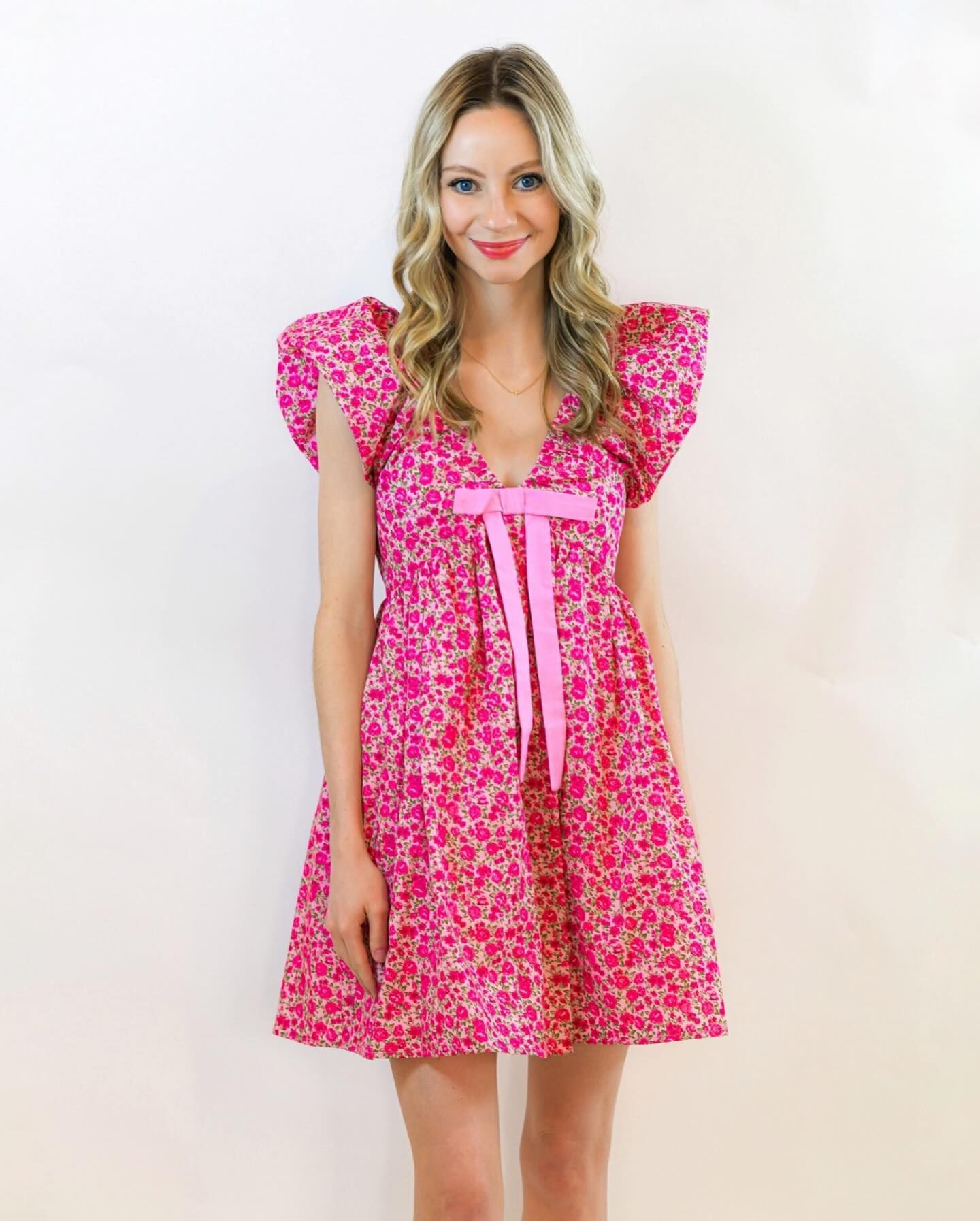 Woman in a pink floral dress with large ruffled sleeves, standing in front of a white wall.