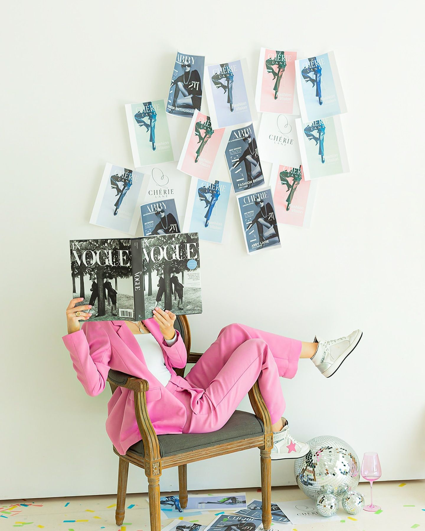 Person in pink suit reading Vogue, feet up, with ballet-themed artwork on white wall.