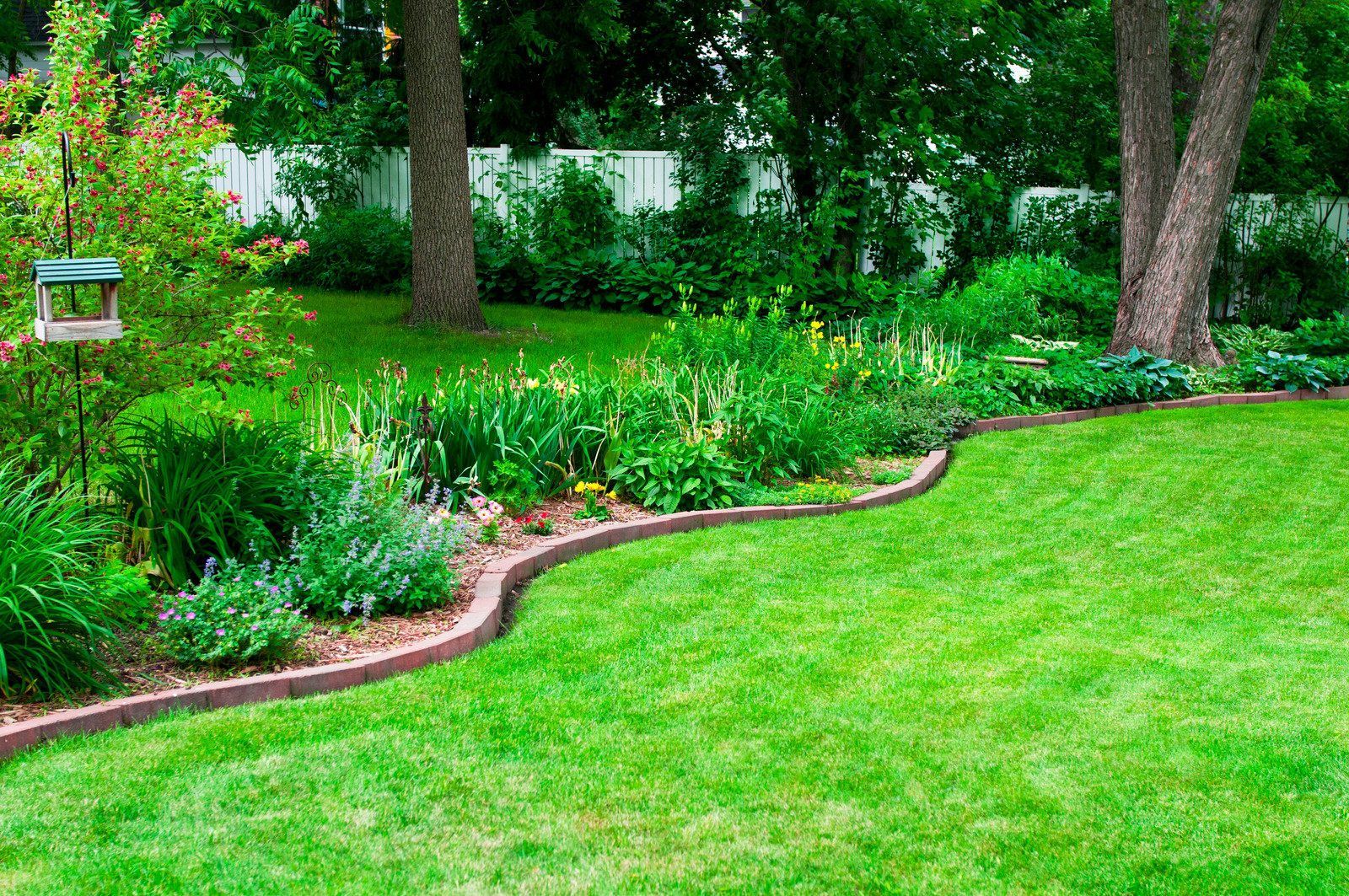 Lush green lawn bordered by a brick edging, leading to a flower bed, with trees and a white fence in the background.