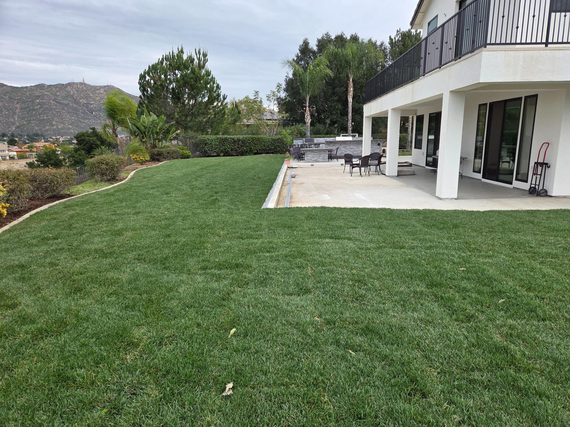 Lush green lawn next to a white house with a patio. Hills in the background. Overcast sky.