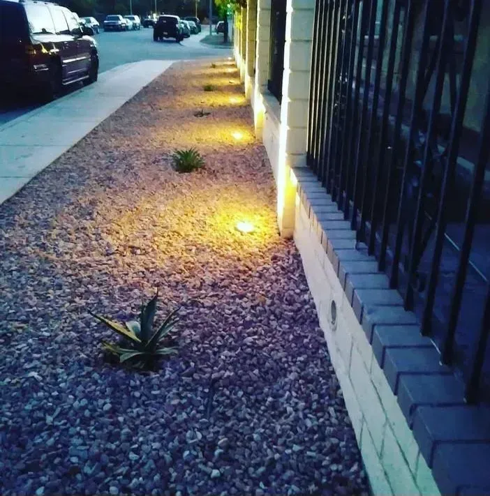 Gravel path along a fence with small plants and lights, next to a street with cars.