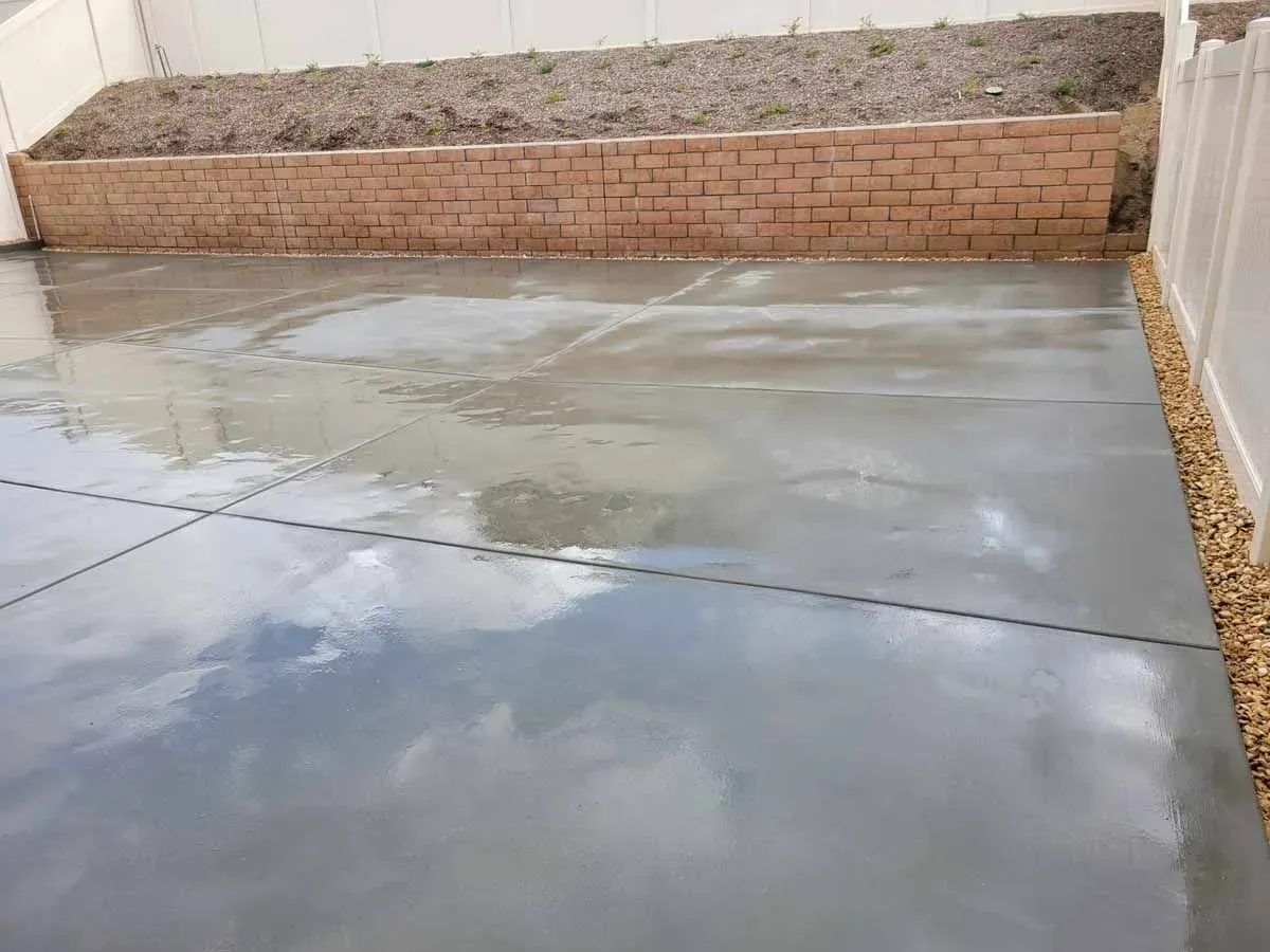 Newly poured gray concrete patio with sky reflection, brown brick retaining wall, and white fence.