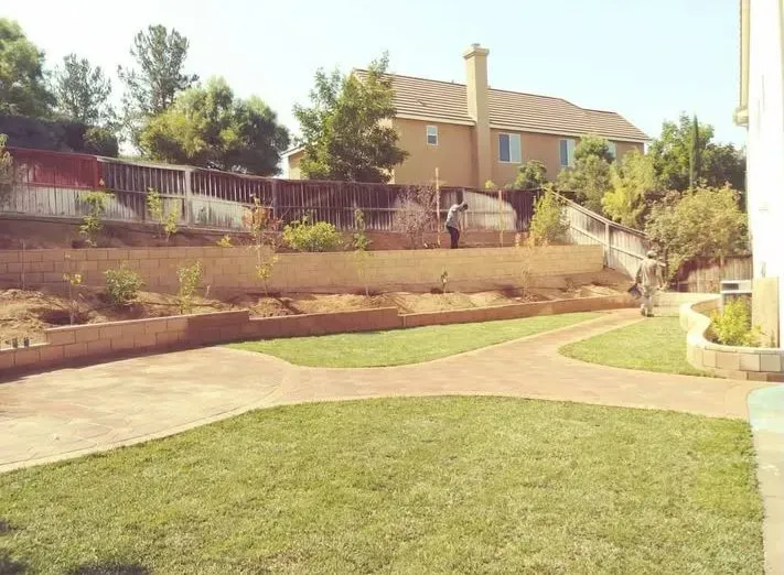 A backyard with grass, a path, and a retaining wall. A person is standing near the top.