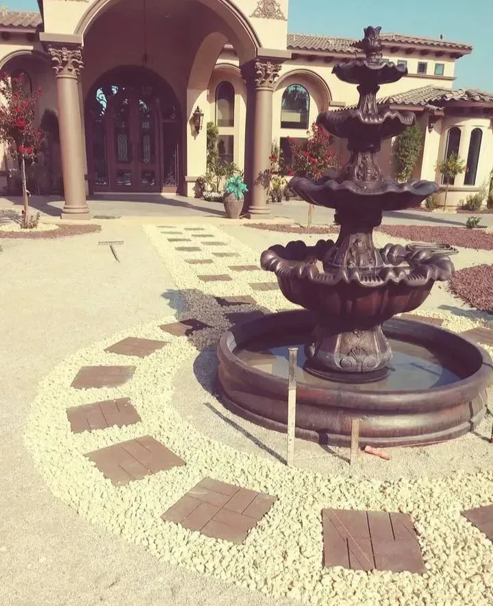 Fountain and walkway leading to an ornate building entrance. Brown fountain, beige gravel, and stone path.