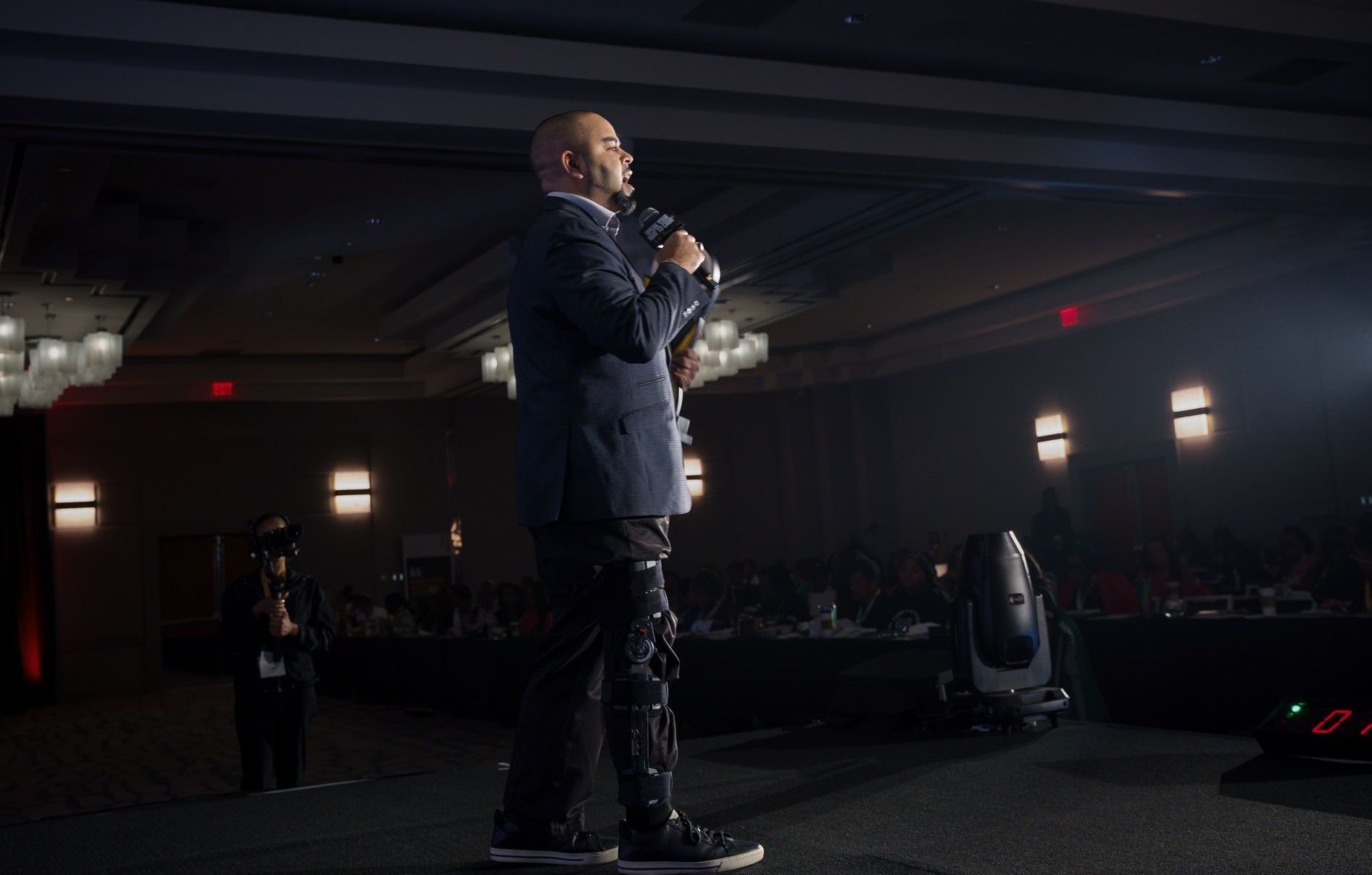 Man in a suit speaks into a microphone on a stage in a dim auditorium.