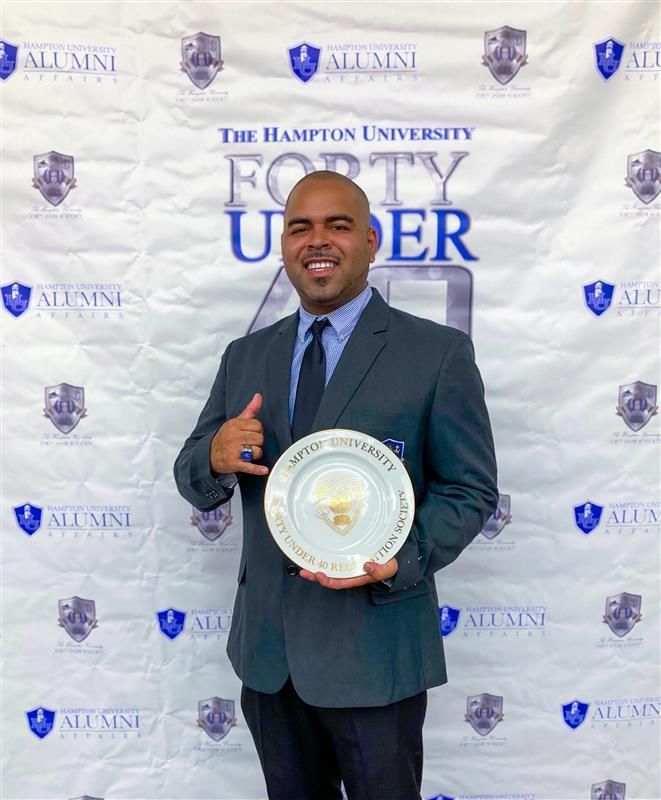 Man in dark suit holding an award plate at Hampton University alumni backdrop