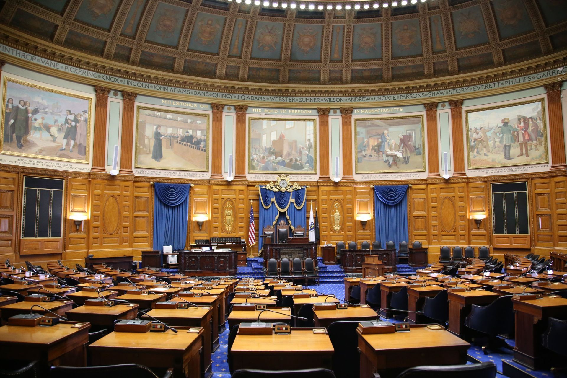 Empty parliamentary chamber with wood desks, blue drapes, and ornate wall paintings