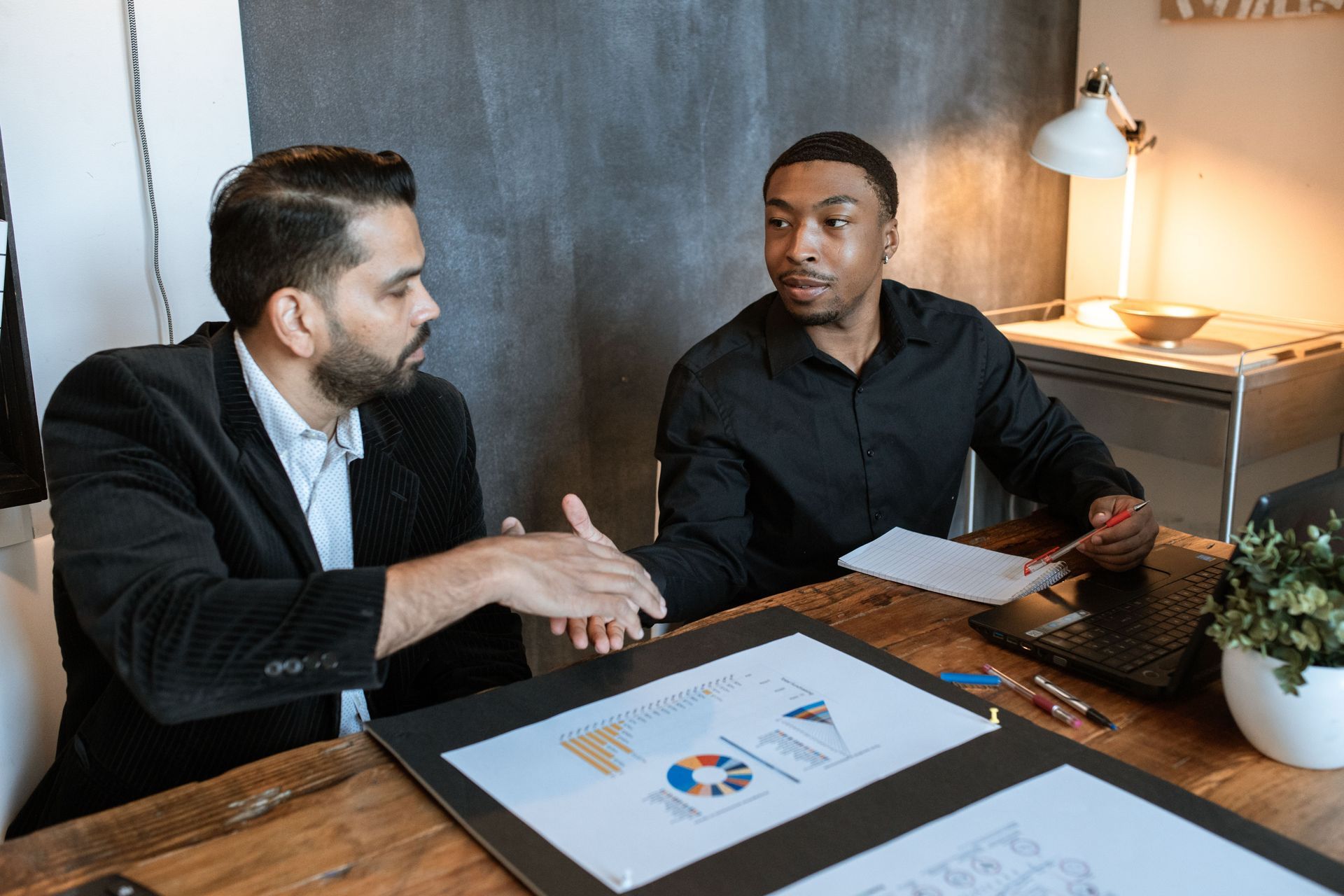 Two professionals in a meeting at a desk with papers, a laptop, and a lamp in a modern office.