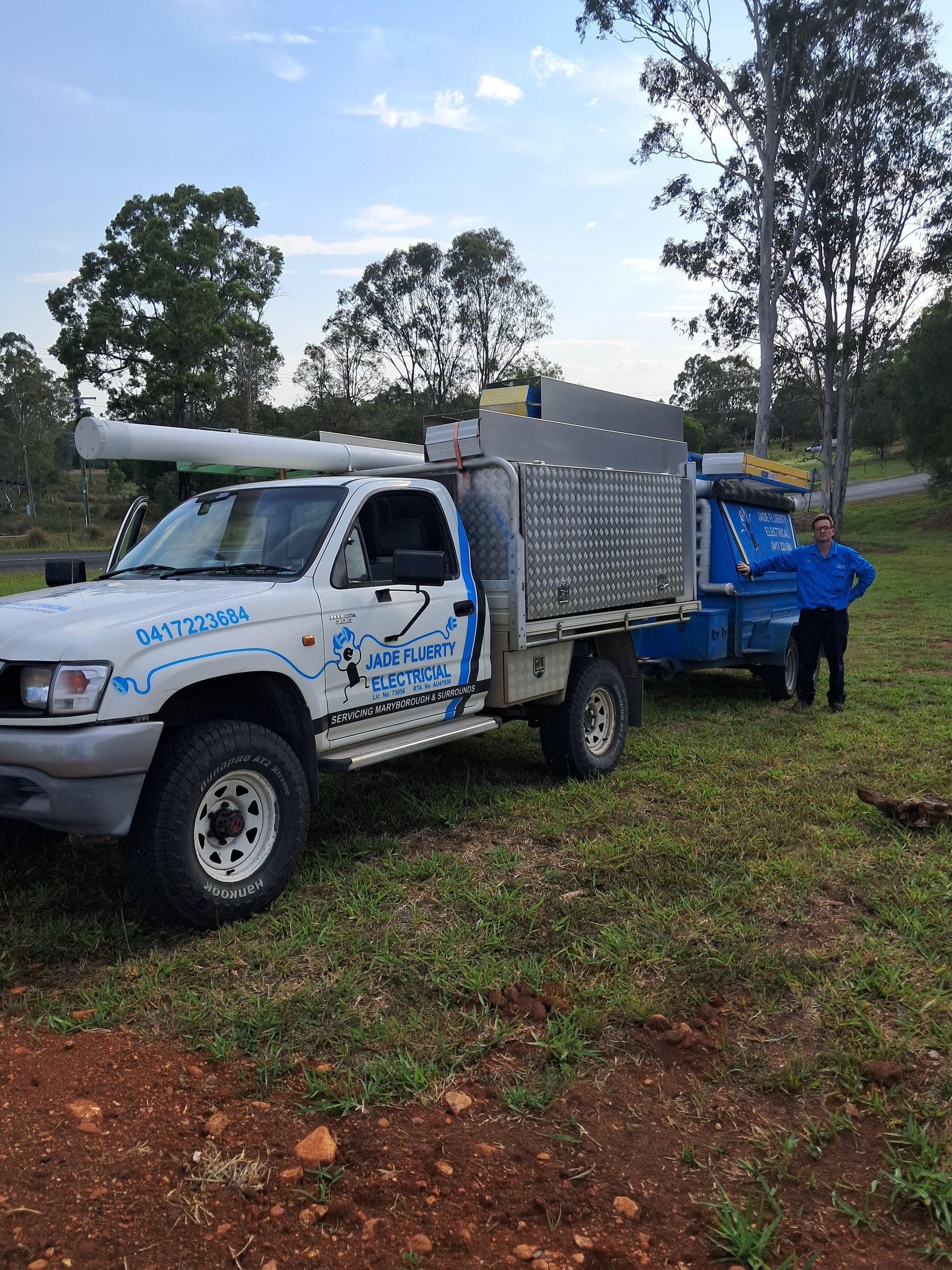 White work truck with trailer; person standing nearby in a grassy area with trees and blue sky in background — Jade Fluerty Electrical In Tinana, QLD