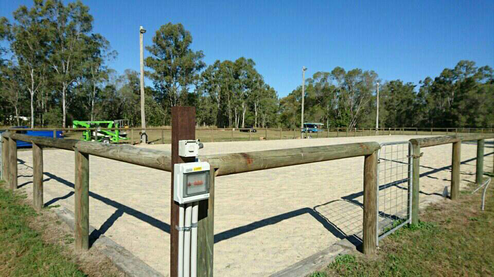 Equestrian arena with wooden fence, sand surface, and tall light poles; trees in the background under blue sky — Jade Fluerty Electrical In Tinana, QLD