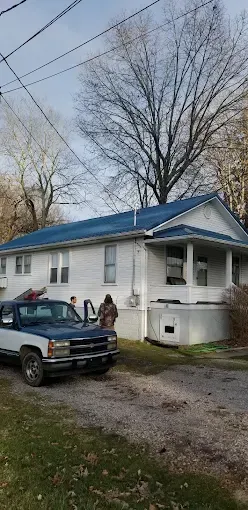 Two people stand near a dark-colored pickup truck parked on a gravel driveway in front of a white house with a blue roof.