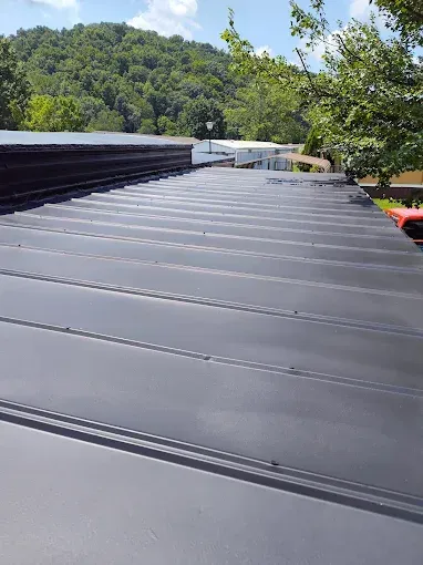 A low-angle view of a matte black standing seam metal roof extending toward a green, tree-covered hillside.