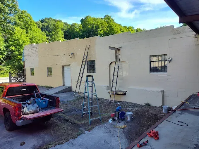 A red truck is parked by a building being painted a light tan color, with ladders leaning against the exterior walls.