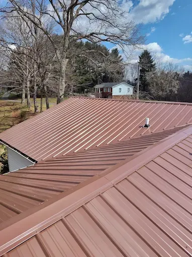 A metal roof with brown standing seam panels, featuring a ridge cap and a small vent pipe, under a blue sky with trees.