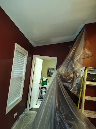A room interior with deep red walls, a white-framed window, and protective plastic sheeting covering an area for painting.