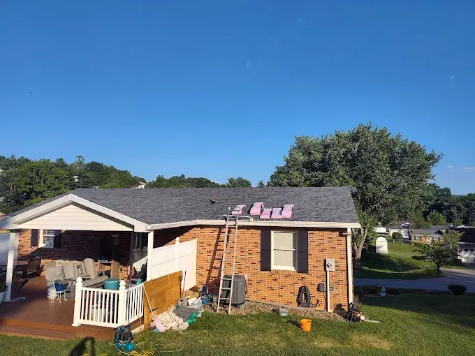 A brick house with a partially shingled roof under repair, featuring a patio, a ladder, and pink underlayment panels.
