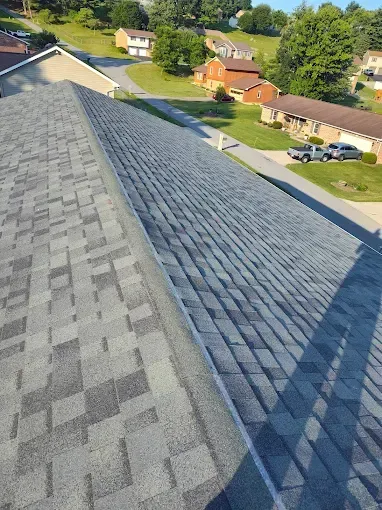 A high-angle view looking down a residential roof with grey shingles toward a suburban street and houses below.