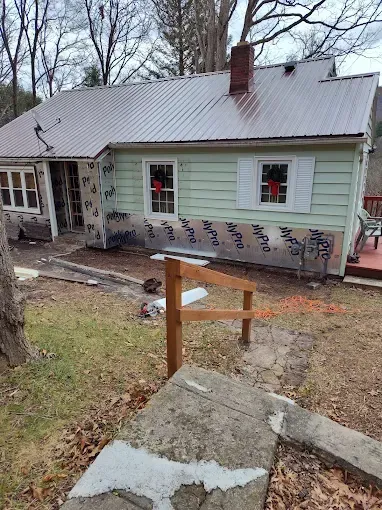 A light green house with a brown metal roof undergoing siding repairs, seen from a concrete path with a wooden railing.