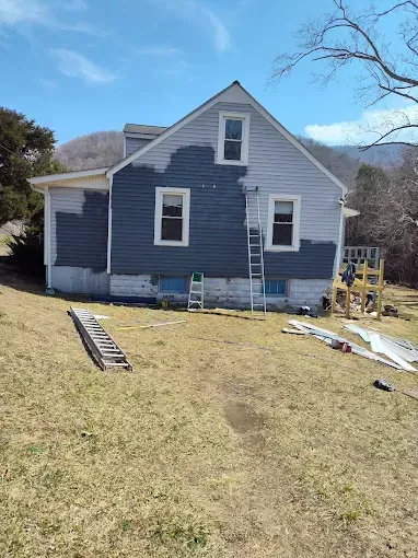 A house exterior under renovation, featuring a partially painted dark gray side wall, ladders, and scaffolding outside.