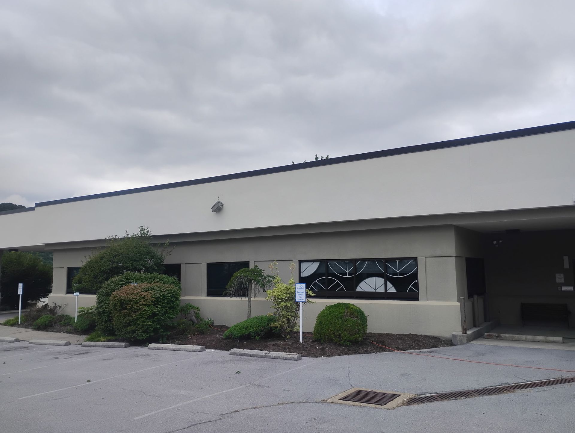 A low-rise, beige commercial building with dark, patterned windows, landscaping, and a parking lot under a cloudy sky.