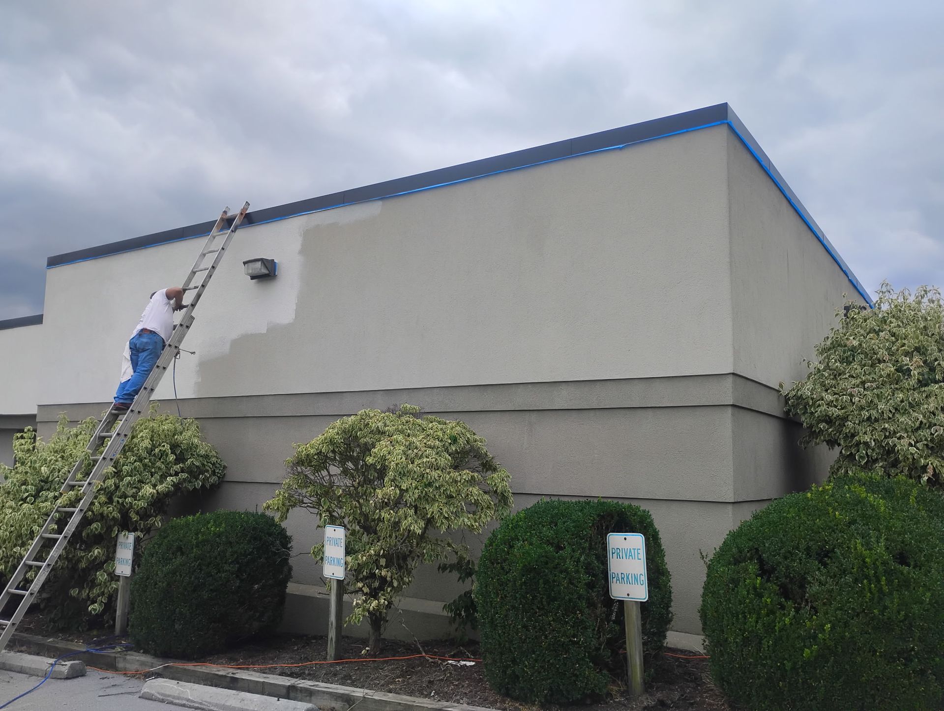 A person standing on an extension ladder paints the upper exterior wall of a commercial building a lighter beige color.
