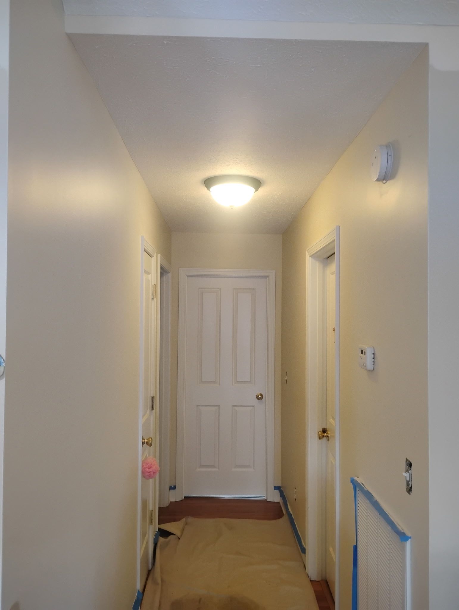 A beige-walled hallway with a lit ceiling light, white doors on the sides and end, and a drop cloth on the floor.