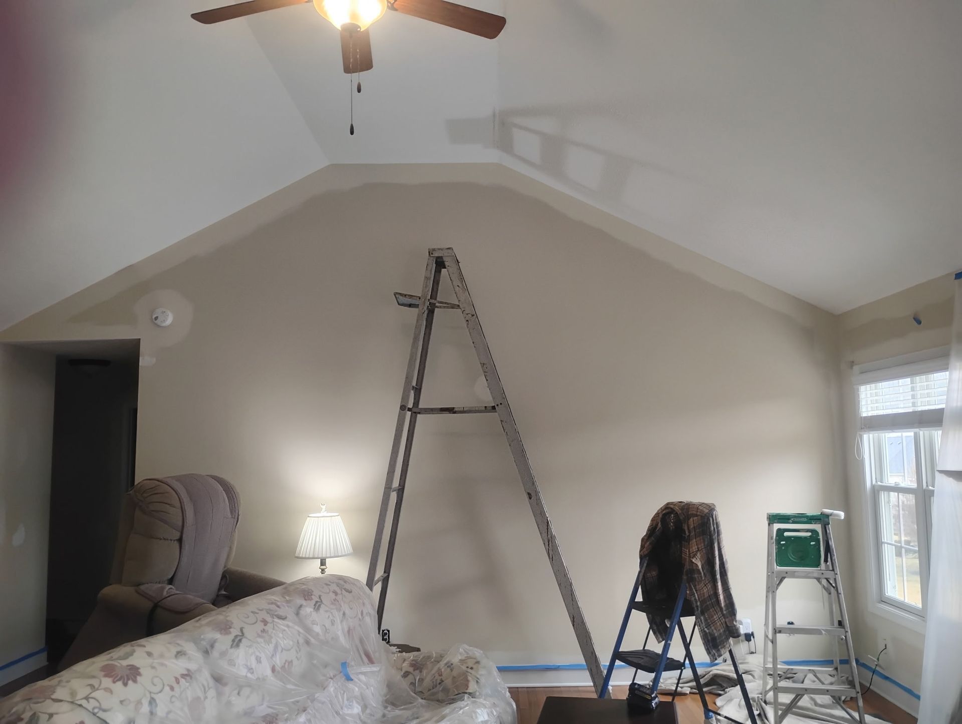 A tall A-frame wall in a living room undergoing renovation, featuring two ladders, a covered couch, and a ceiling fan.