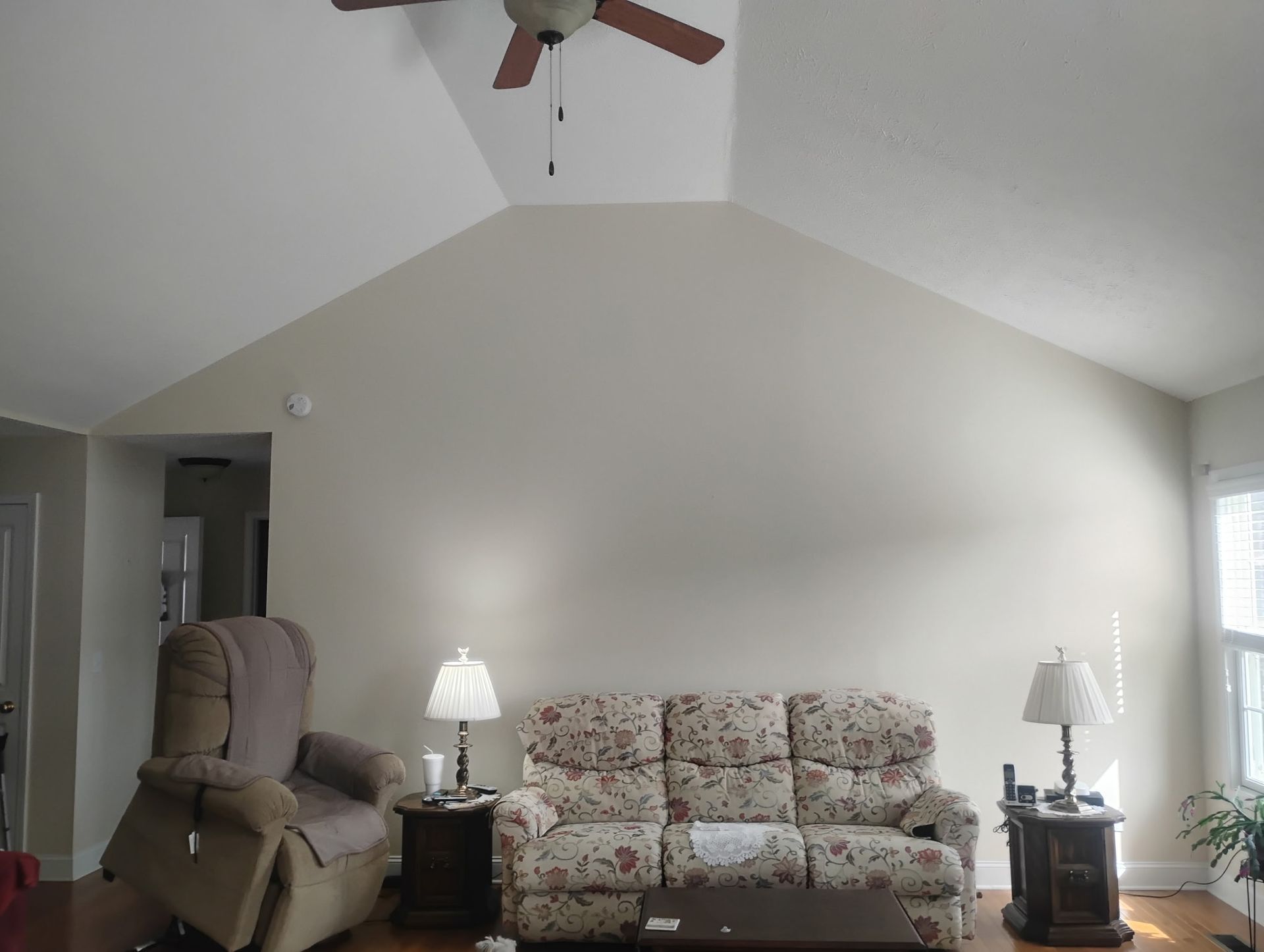 A living room featuring a tall, vaulted ceiling, a beige floral sofa, a matching recliner, and two side tables with lamps.