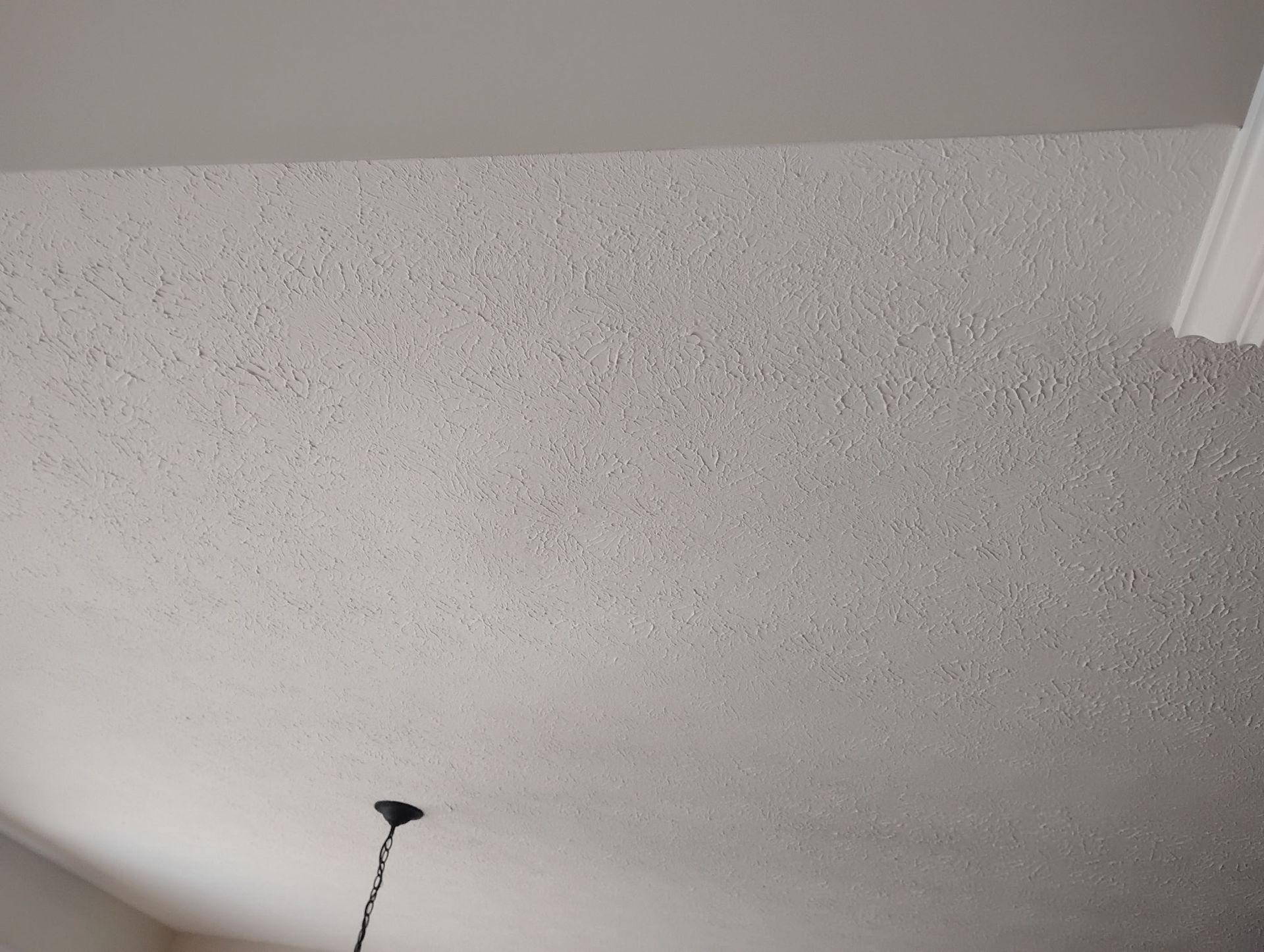 A close-up view of a white, textured popcorn ceiling with a small portion of a light fixture hanging in the corner.