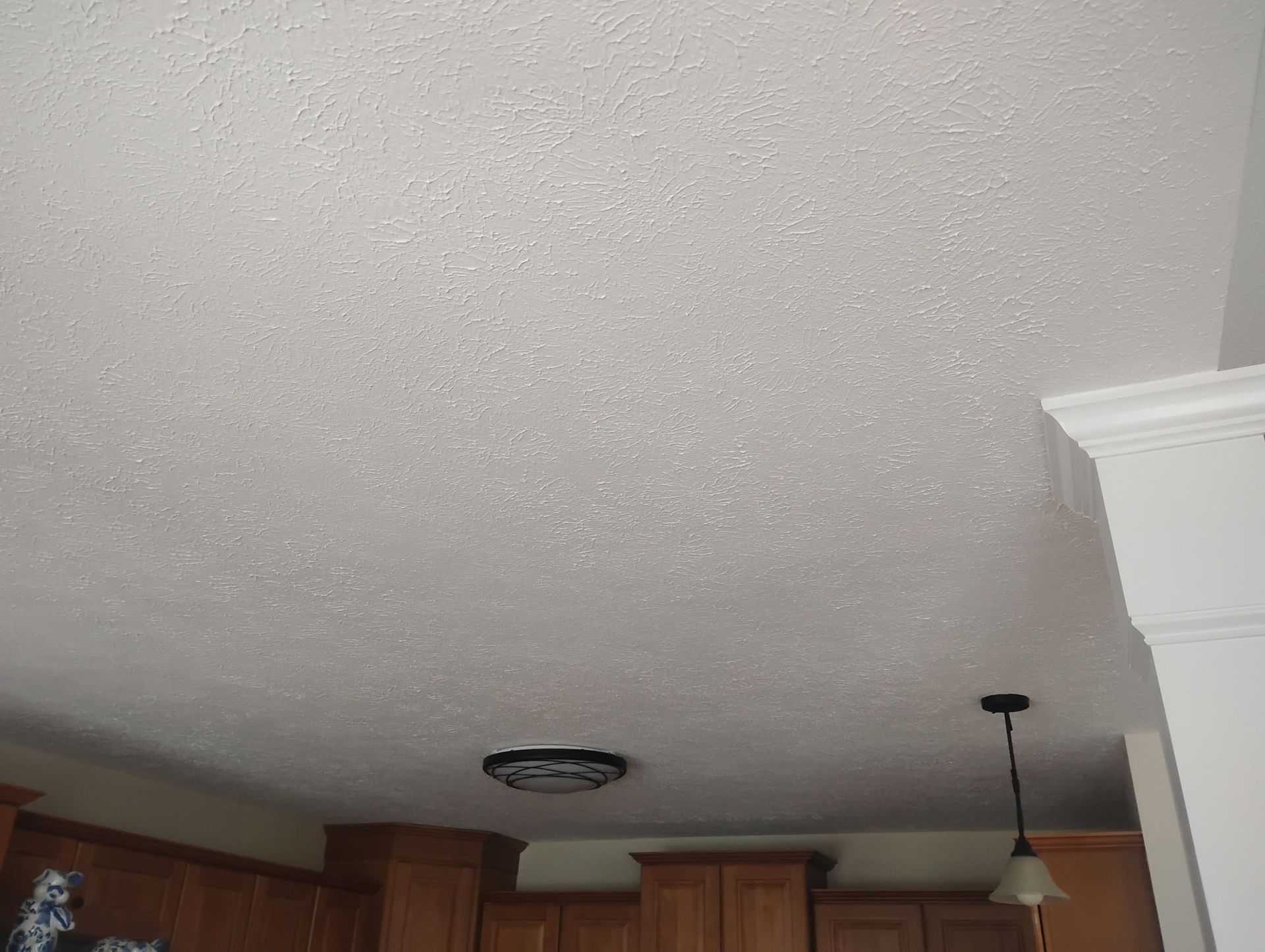 A white, textured ceiling with a circular flush-mount light fixture and a pendant light hanging over wooden cabinets.