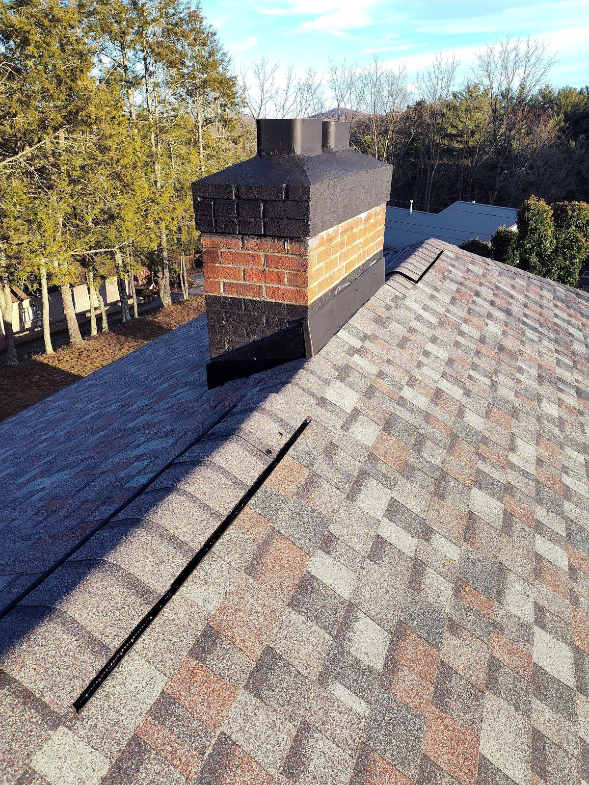 A chimney with a brick middle and painted black base and top, protruding from a shingled roof with a black roof vent strip.